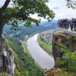 Mehrere Menschen rechts im Bild schauen von einer aus der Anhöhe herausragenden Plattform auf das Elbtal in der Mitte und auf aufragende Felsen des Elbsandgebirges am linken Rand des Bildes. Links im Vordergrund ein Baum mit Blättern.